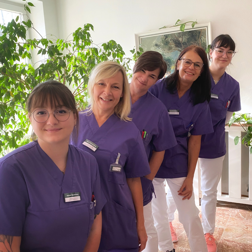 Foto des Teams des medizinischen Funktionsdienstes der Klinik Wartenberg in ihren lilanen Kasacks. Die Gruppe steht hintereinander aufgereiht und schaut in die Kamera. Im Hintergrund erkennt man verschiedene Pflanzen und die Sonne scheint durch das Fenster hinein.