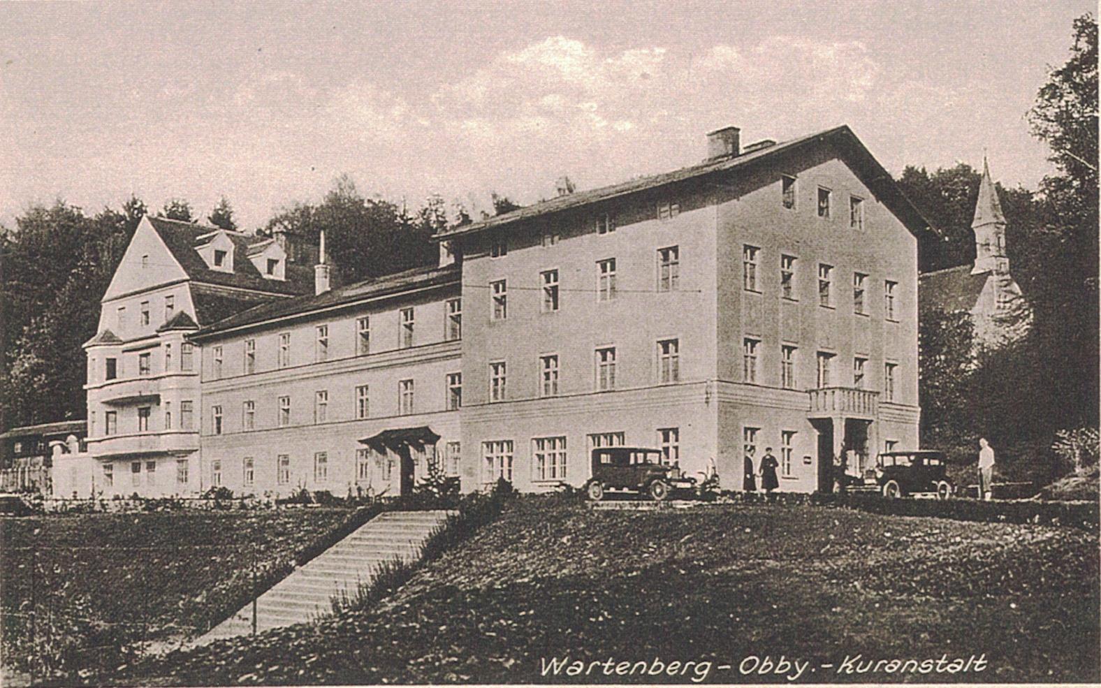 Historische schwarz-weiß Aufnahme vom Altbau der Klinik Wartenberg. Im Hintergrund erkennt man die alte Bründlkirche, die heute nicht mehr existiert. Vor dem Gebäude stehen Fahrzeuge und Personen.