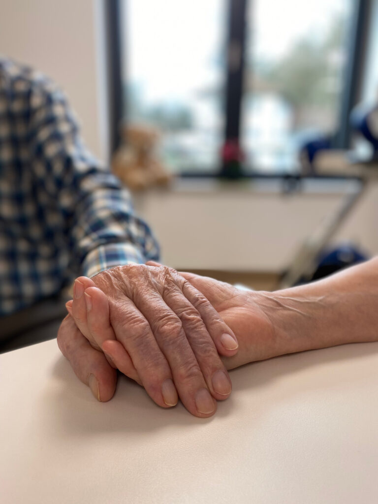 Auf dem Foto sieht man zwei Hände. Die untere Hand einer Frau hält die obere Hand eines älteren Patienten, den man teilweise unscharf im Hintergrund erkennen kann. Im Hintergrund sieht man außerdem ein Fenster, durch das man in den Innenhof der Klinik Wartenberg schauen kann.