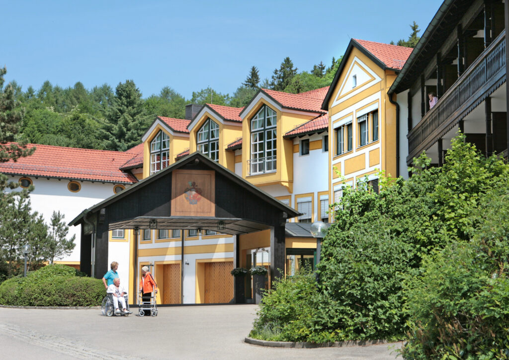 Auf dem Foto sieht man den Haupteingang der Klinik Wartenberg mit der hölzernen Überdachung. Davor stehen zwei Patientinnen und eine Patientenbetreuerin. Rechts im Bild sieht man ein grünes Gebüsch. Im Hintergrund steht das Hauptgebäude. Dahinter erkennt man die Bäume des Klinikwaldes. Der Himmel ist blau, es scheint ein sommerlicher Tag zu sein.