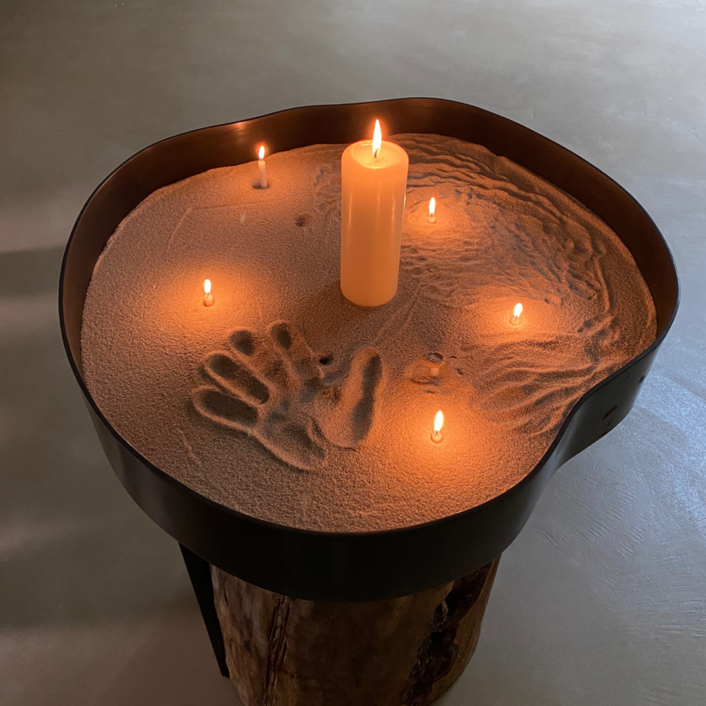 Auf dem Foto sieht man ein Objekt, das im Raum der Stille im Neubau der Klinik Wartenberg steht. Dieses wurde aus einem Baumstamm gefertigt, auf dem in einer Metallschale Sand liegt. In dem Sand stecken mehrere Kerzen und jemand hat einen Handabdruck hinterlassen.