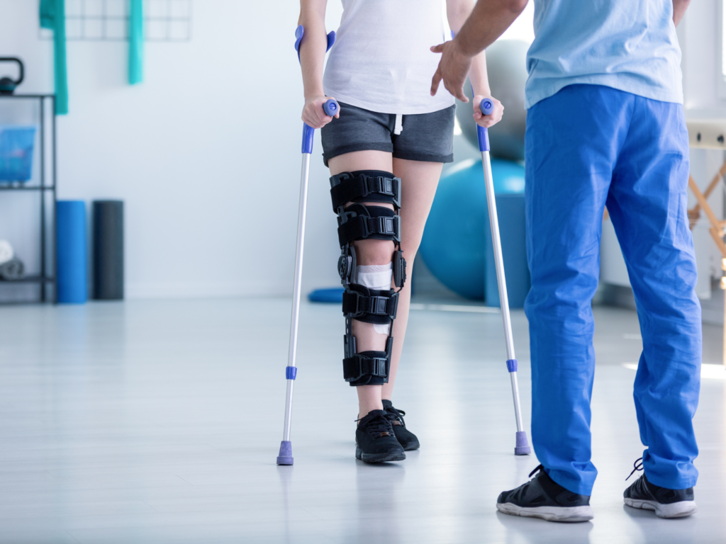 Auf dem Foto sieht man eine Patientin mit einer Beinschiene und Unterarmgehstützen in einem Gymnastiksaal. Ihr gegenüber steht ein Physiotherapeut, der sie beim Gehen anleitet. Im Hintergrund erkennt man weitere Geräte und Utensilien aus der Physiotherapie.