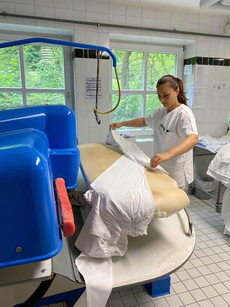 Auf dem Foto sieht man eine Mitarbeiterin in der Wäscherei der Klinik Wartenberg, wie sie an einer Maschine arbeitet und Wäsche vorbereitet.