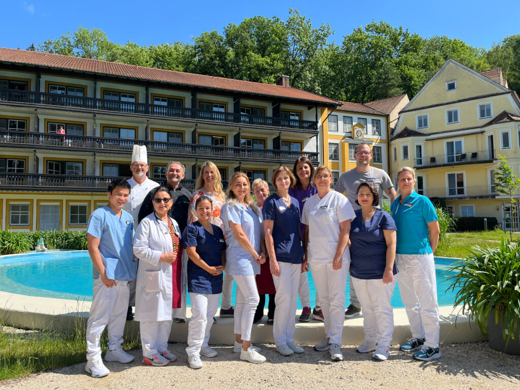 Auf dem Foto sieht man eine Gruppe von Mitarbeitenden verschiedener Berufsgruppen der Klinik Wartenberg vor dem Springbrunnen im Innenhof der Klinik stehen. Das Foto wurde im Rahmen der „Klinik-Familie“ aufgenommen, um die Vielfalt der Klinik Wartenberg zu zeigen. Im Hintergrund erkennt man das Klinikgebäude sowie den Wald. Der Himmel ist blau und die Sonne scheint, es scheint ein sommerlicher Tag zu sein.