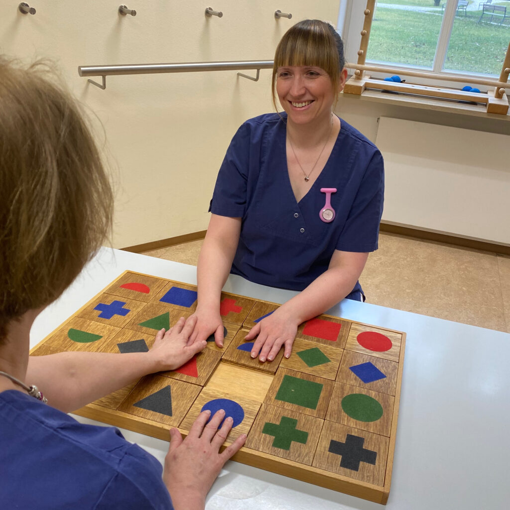 Auf dem Foto sieht man eine Ergotherapeutin, die mit einer Patientin ein Spiel an einem Tisch spielt. Die beiden sitzen im Therapieraum der Ergotherapie, im Hintergrund erkennt man weitere Utensilien der Ergotherapie, sowie ein Fenster, durch das man in den Innenhof schauen kann.