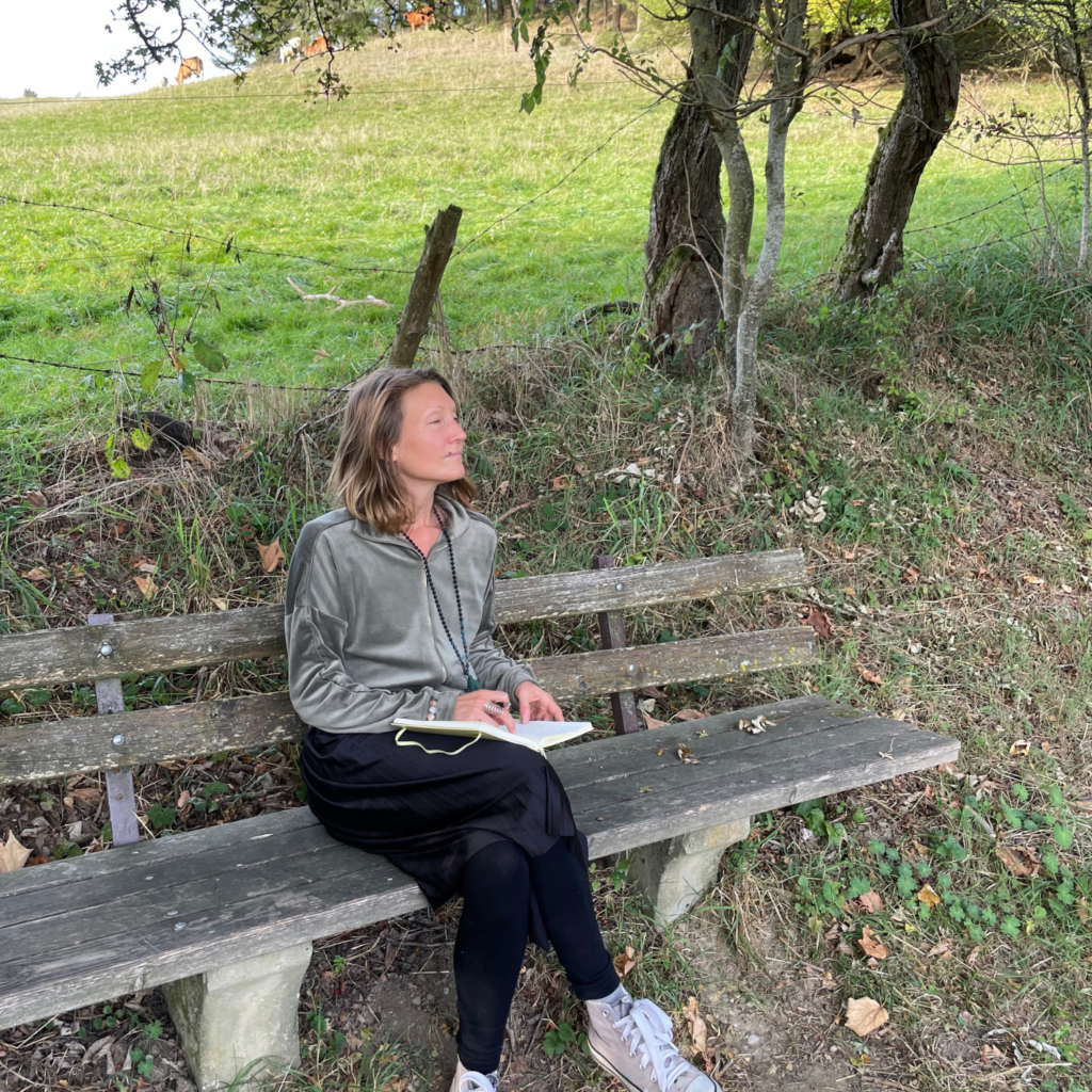Bild von Britta Jörg die auf einer Bank sitzt und etwas in ein Buch schreibt. Im Hintergrund Stacheldraht und eine große Wiese