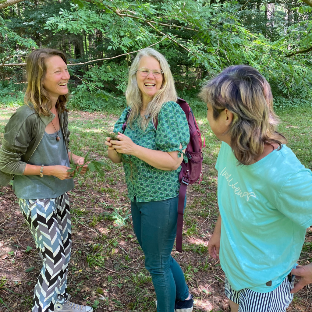 Auf dem Foto sieht man drei Frauen, die im Wald stehen und sich gegenseitig anlachen.
