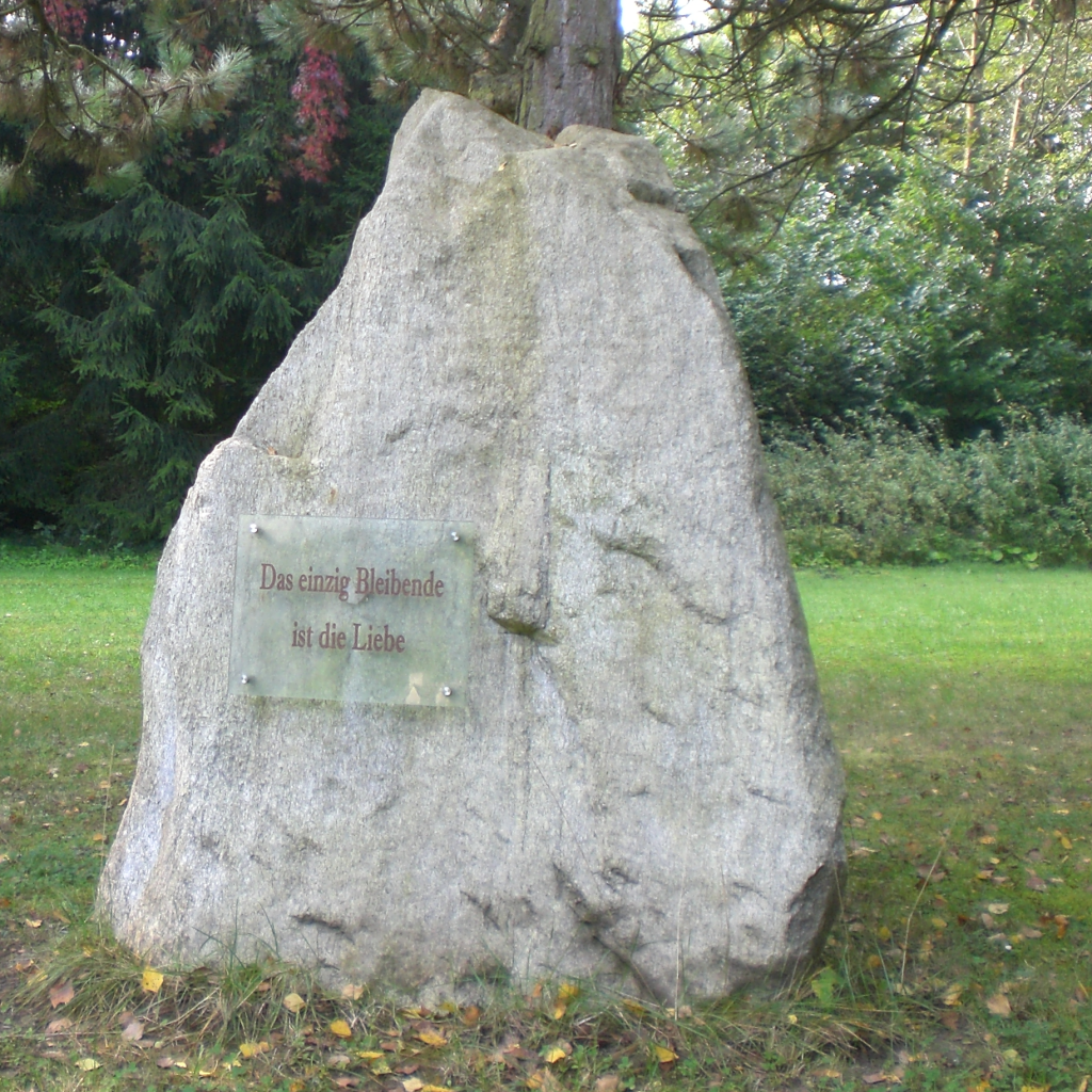 Bild eines großen Steins, der auf der Wiese steht mit einem Schild "Das einzige Bleibende ist die Liebe" Im Hintergrund Bäume und Sträucher.