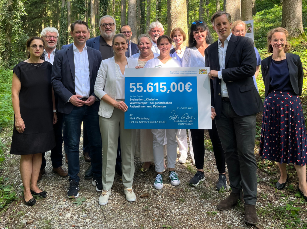 Übergabe der Förderung der Waldtherapie. 55.615€ für das Projekt "Evalution Klinische Waldtherapie bei geriatrischen Patientinnen und Patienten". 2 Personen halten die Förderung in der Hand, dahinter 13 Personen in dem Klinik Wald.