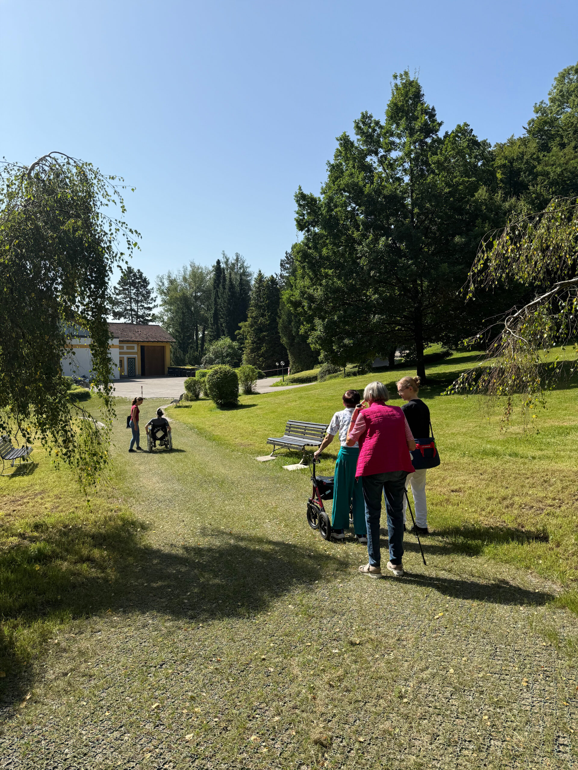 Eine Gruppe von Patientinnen und Patienten, teilweise mit Hilfsmitteln wie Rollatoren und Rollstühlen, gehen gemeinsam mit den Therapeutinnen den Weg von Waldtherapiepfad hinunter zurück zur Klinik. Man sieht im Hintergrund das Gebäude. Der Weg ist befestigt, rechts und links sieht man das grüne Gras und seitlich am Wegesrand steht jeweils eine Bank. Außen sieht man die Bäume auf dem Klinikgelände stehen. Der Himmel ist blau und es sieht nach einem sommerlichen Tag aus.