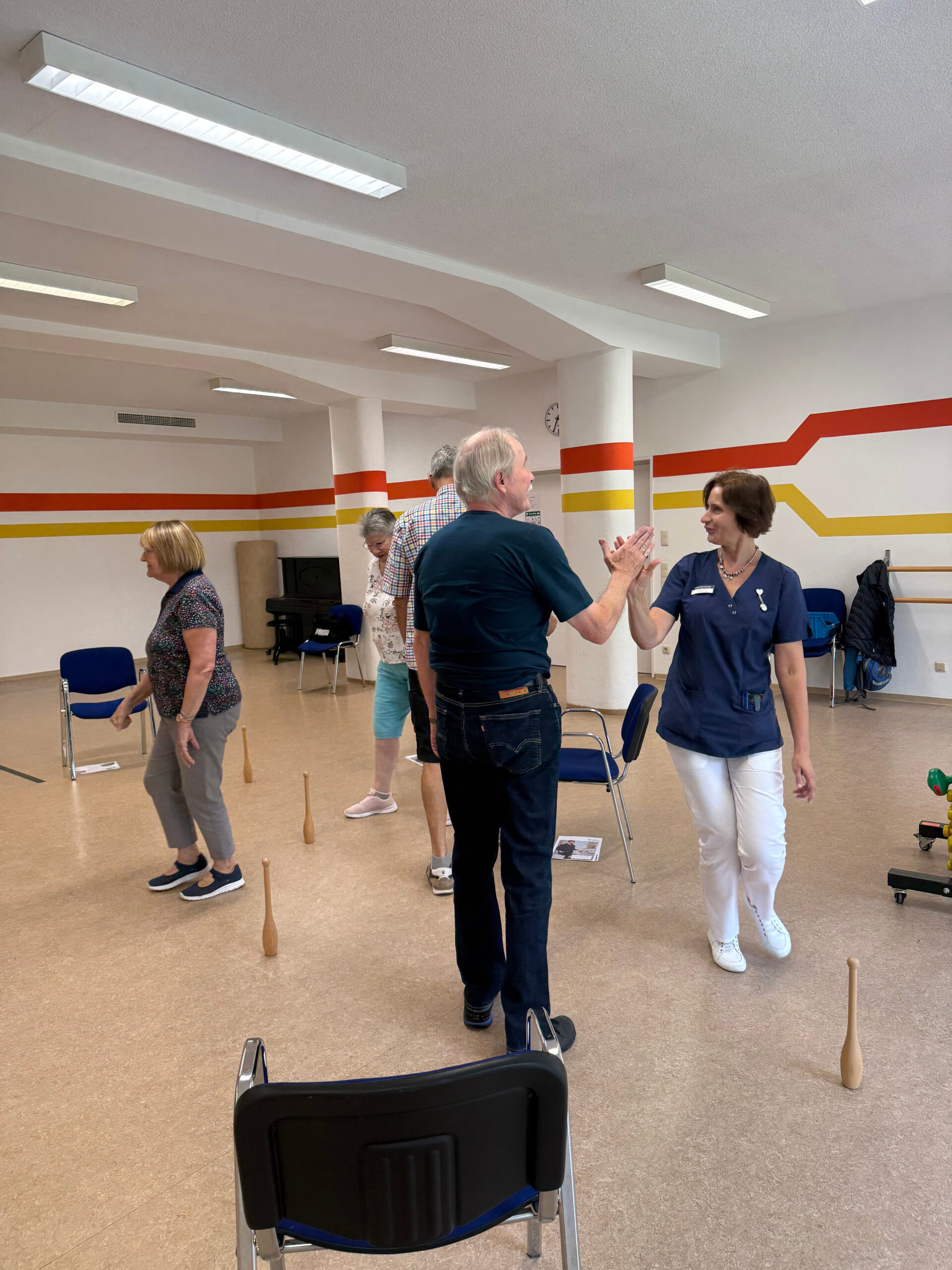 Auf dem Foto sieht man eine Gruppe von Teilnehmenden des MoSi Kurses 2025 zusammen mit der Therapeutin im Gymnastiksaal der Klinik Wartenberg. Ein Teilnehmer, der im Vordergrund zu sehen ist, klatscht seine Hand mit der Therapeutin ab. Die Gruppe führt Übungen in Bewegung durch. Im Raum stehen Stühlen und andere Hilfsmittel.