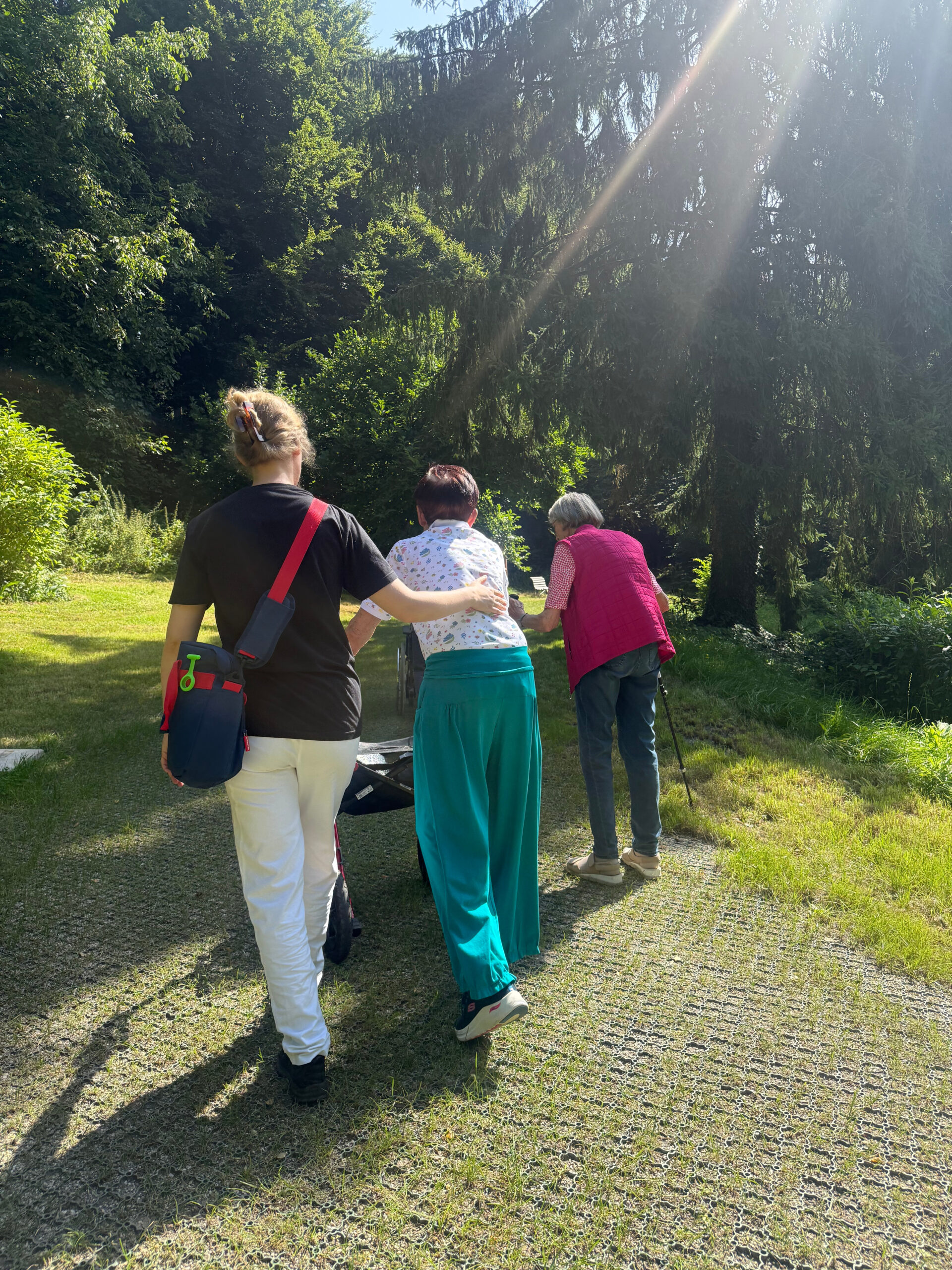 Zwei Patientinnen gehen gemeinsam mit der Therapeutin den Weg zum Waldtherapiepfad hoch. Die Sonnenstrahlen scheinen von rechts oben durch die Bäume ins Bild. Der Weg ist befestigt, rechts und links sieht man das grüne Gras, weiter hinten die grünen Bäume.