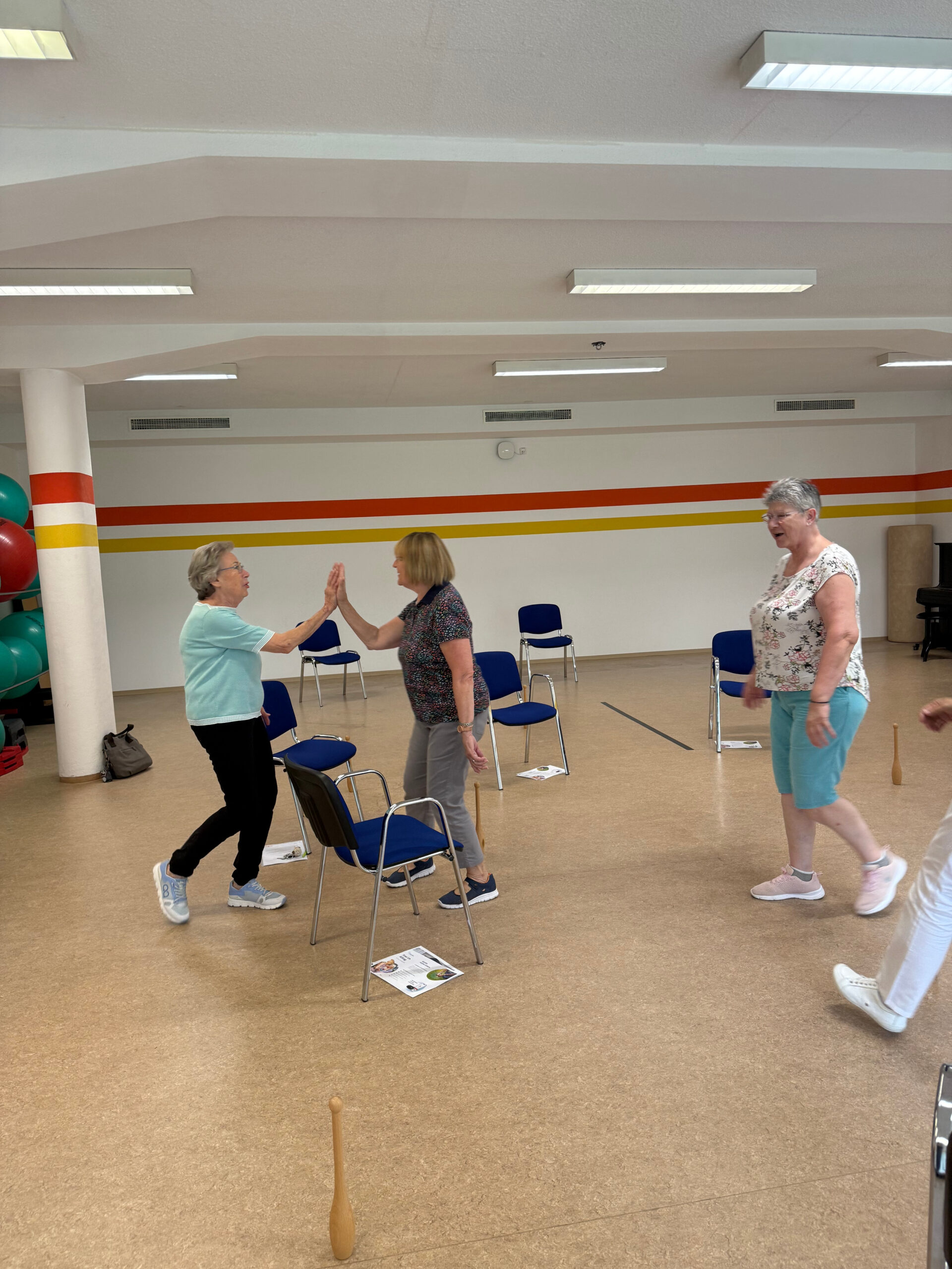 Auf dem Foto sieht man drei Teilnehmerinnen des MoSi-Kurses 2025. Zwei von ihnen klatschen sich gegenseitig mit den Händen ab. Die Gruppe befindet sich im Gymnastiksaal der Klinik Wartenberg. Im Raum stehen Stühle und im Hintergrund sieht man Gymnastikbälle.