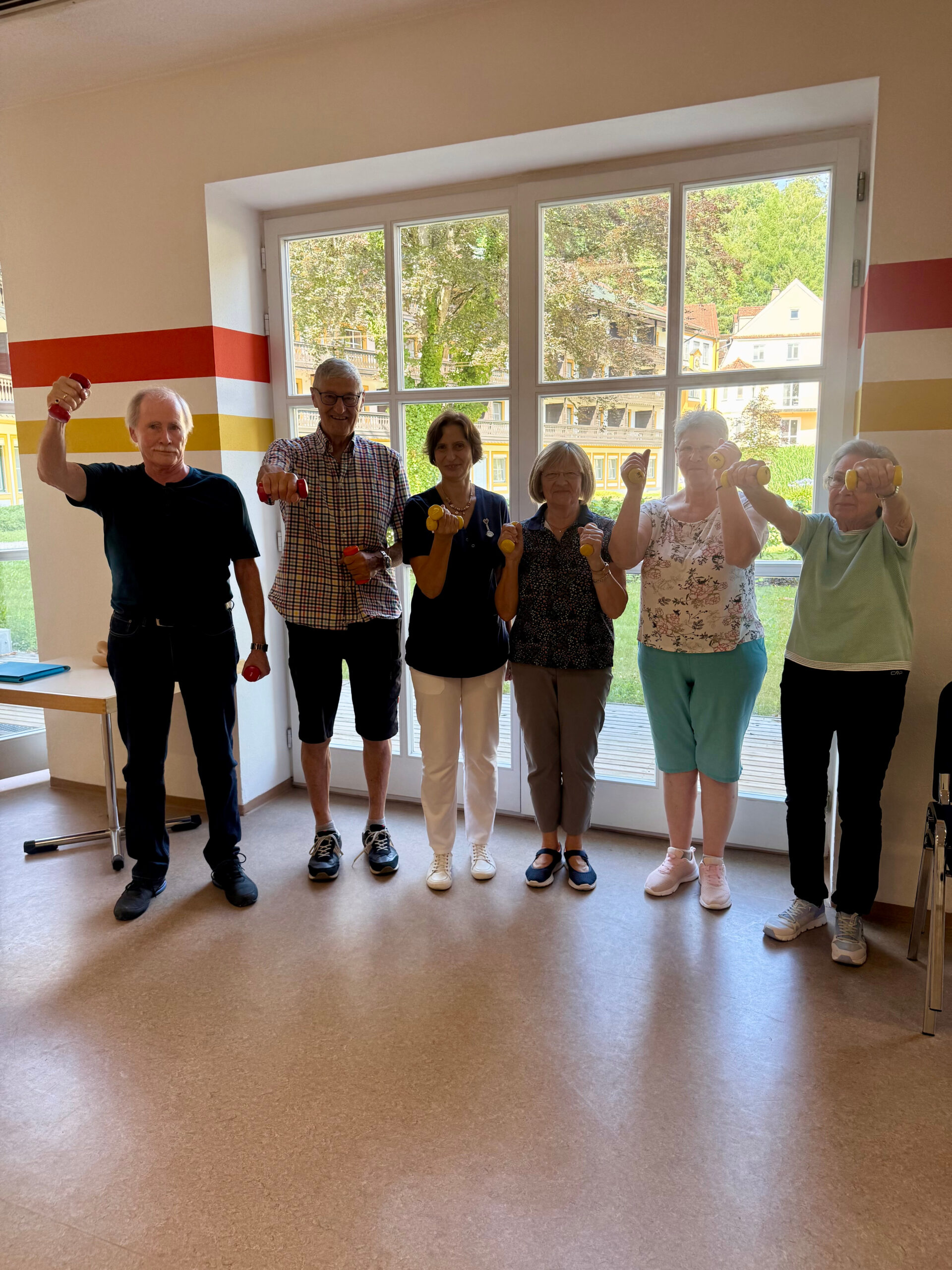 Eine Gruppe von Teilnehmenden des MoSi-Kurses posiert im Gymnastiksaal der Klinik Wartenberg. Sie halten kleine Kurzhanteln in die Höhe. Im Hintergrund sieht man den grünen Innenhof der Klinik Wartenberg.