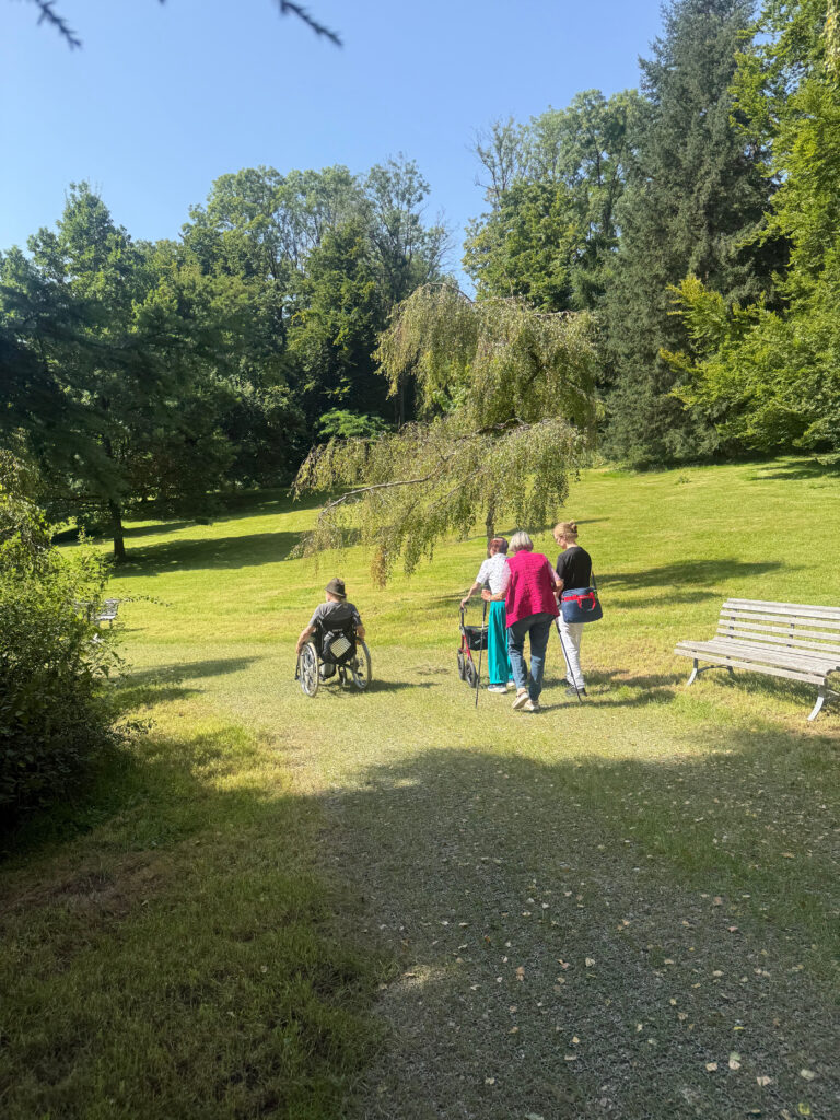 Eine kleine Gruppe von Patienten geht den Waldtherapiepfad hinunter zurück zur Klinik. Der Rasen im Hintergrund ist grün und man sieht die Bäume des Waldes. Der Himmel ist blau. Es ist ein sommerlicher Tag.