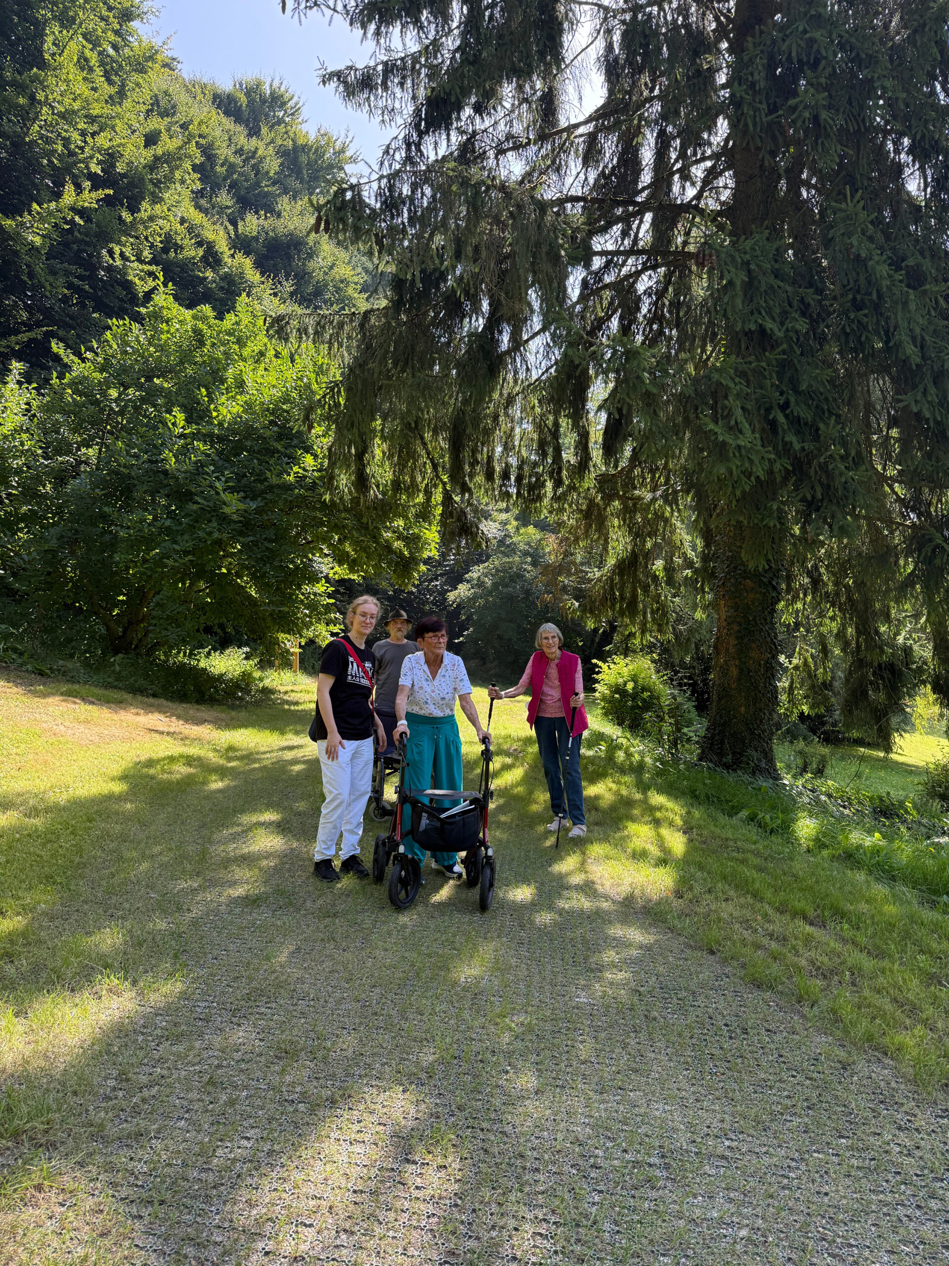 Eine kleine Gruppe von Patienten steht zusammen mit der Waldtherapeutin auf dem Waldtherapiepfad. Der Weg ist befestigt, an den Seiten sieht man den grünen Rasen und im Hintergrund die Bäume des Waldes.