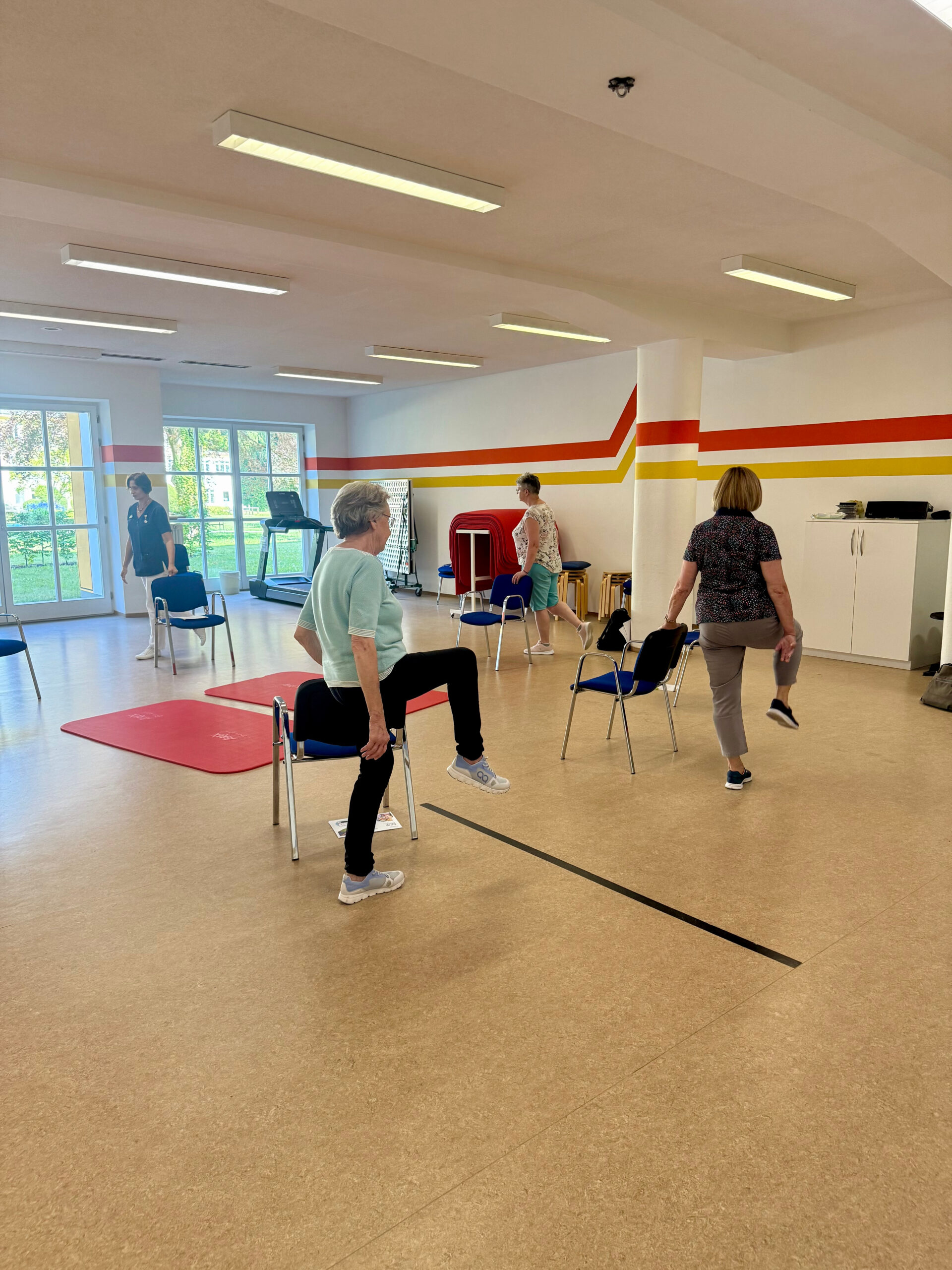 Patientinnen und Therapeutin im Gymnastiksaal der Klinik Wartenberg. Führen Übungen hinter ihren Stühlen durch, die im Kreis aufgestellt sind. Im Hintergrund sieht man den grünen Innenhof der Klinik.