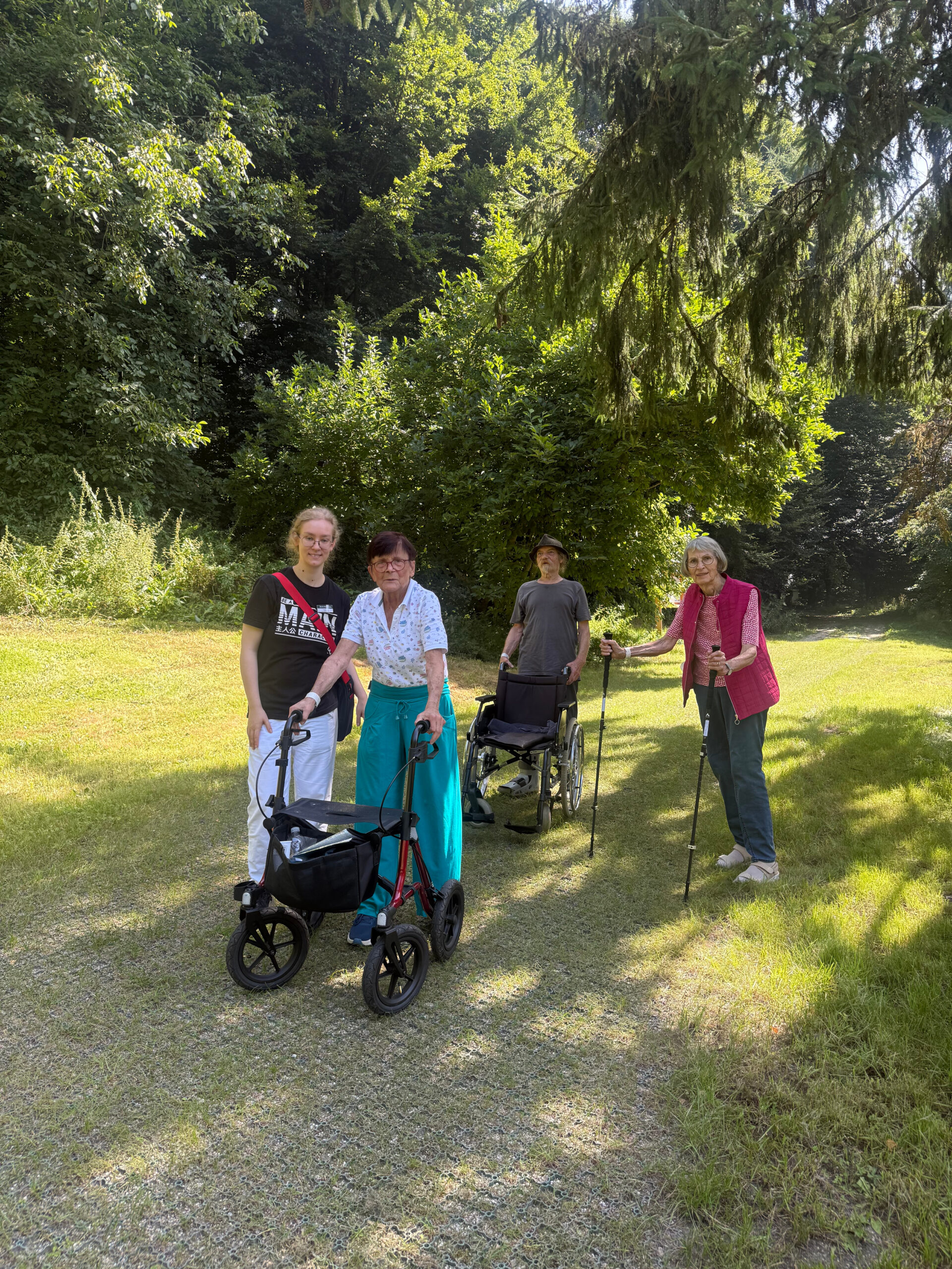 Die teilnehmenden Patientinnen und Patienten der Waldtherapie posieren gemeinsam mit der Waldtherapeutin auf dem zertifizierten Waldtherapiepfad für das Foto. Sie stehen auf dem befestigten Weg, an den Seiten im Hintergrund sieht man das grüne Gras und die Bäume vom Wald. Die Sonne scheint.