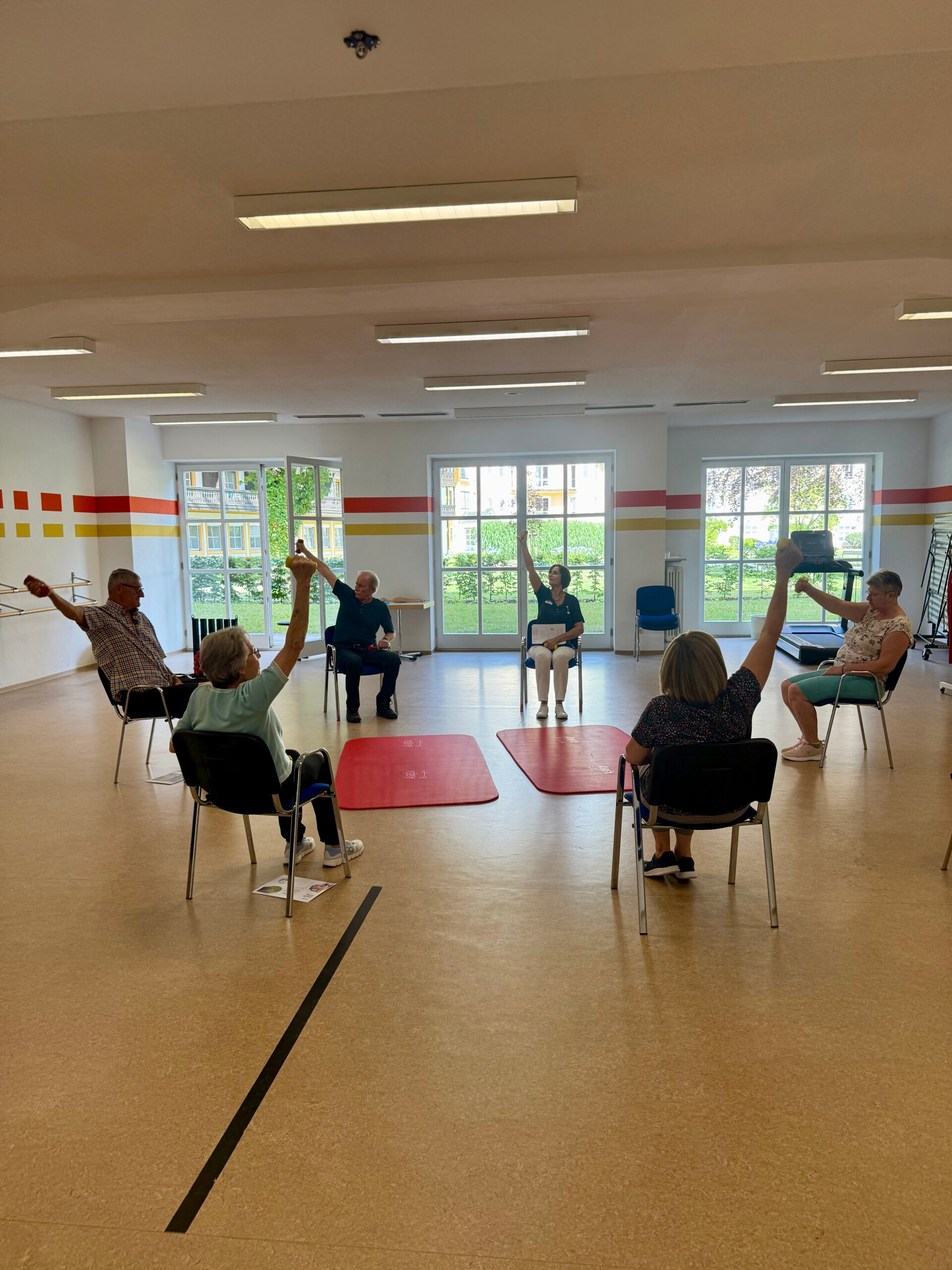 Eine Gruppe von Teilnehmenden am MoSi-Kurs sitzt in einem Stuhlkreis im Gymnastiksaal der Klinik Wartenberg und führt unter Anleitung der Therapeutin eine Übung durch, bei der jeder einen Arm mit einer Kurzhantel in die Höhe hält. Im Hintergrund sieht man den grünen Innenhof der Klinik.