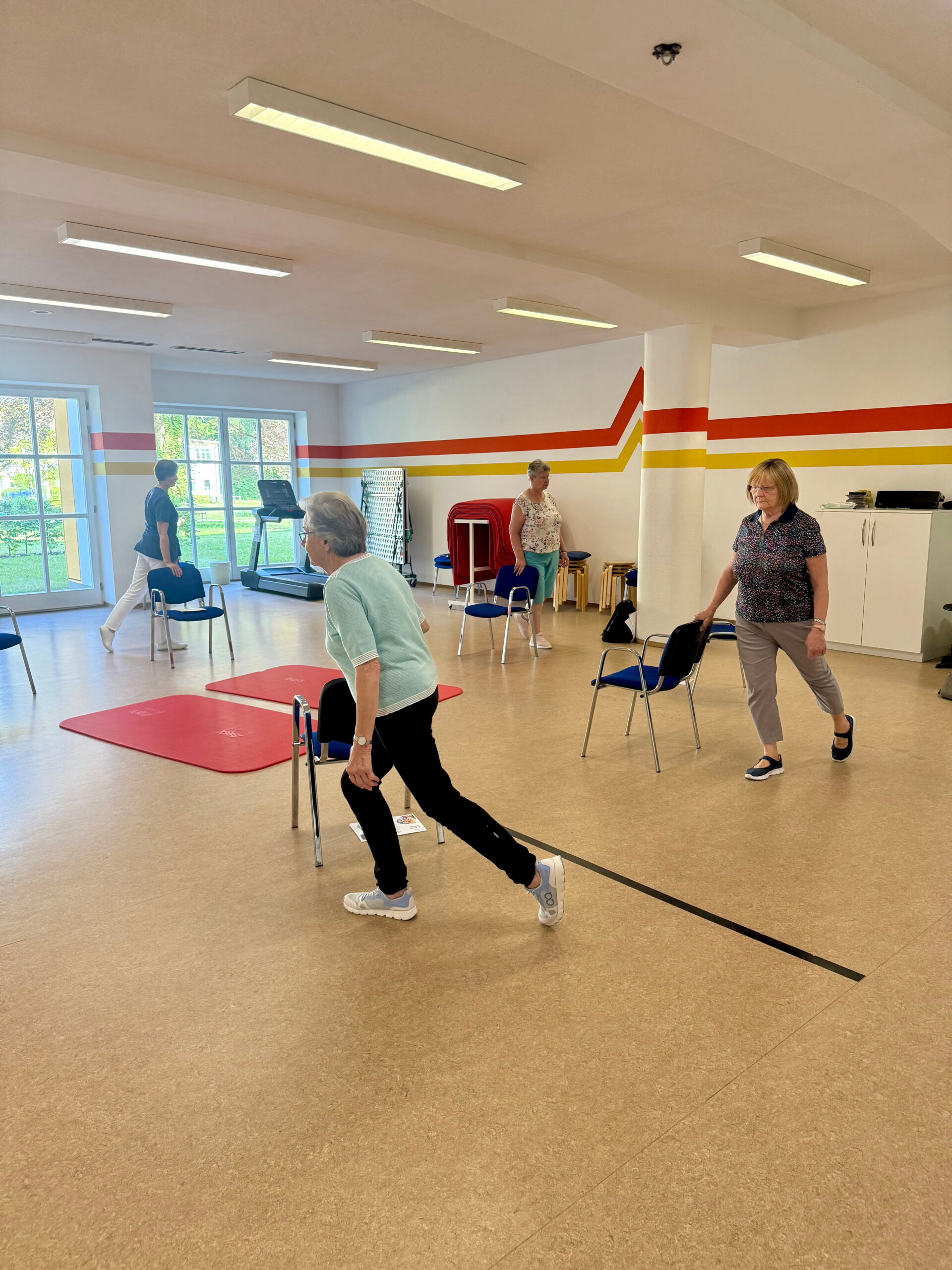 Die Teilnehmenden des MoSi-Kurses führen gemeinsam mit der Therapeutin Übungen in einem Stuhlkreis durch. Jeder steht hinter seinem Stuhl und streckt ein Bein nach hinten. Die Gruppe befindet sich im Gymnastiksaal der Klinik Wartenberg. Im Hintergrund sieht man den grünen Innenhof der Klinik.